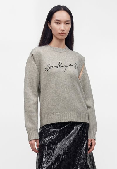 Femme aux longs cheveux noirs porte un pull gris avec une inscription noire, une découpe à l'épaule et une jupe noire brillante texturée sur un fond uni.