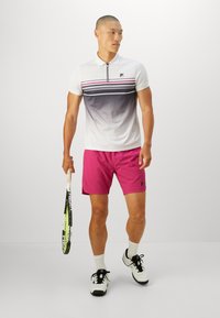 White polo shirt with gray ombre and striped accents, paired with pink shorts. Holding a tennis racket, wearing black and white shoes.