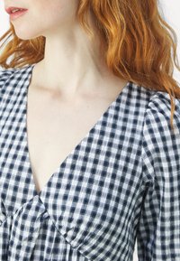 Personne aux cheveux roux portant un haut à col en V à carreaux noir et blanc avec des manches longues, visible du cou au haut du torse.