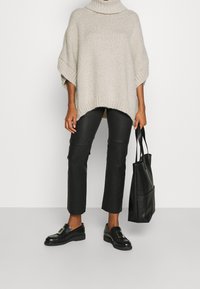 Beige gestrickter Poncho mit hohem Kragen, lockeren Ärmeln und geripptem Saum, kombiniert mit schwarzen Lederhosen und schwarzen Slippern. Tragetasche inklusive.