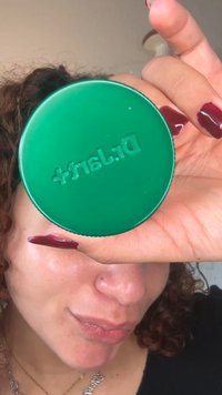Grønn rund beholder merket "Dr. Jart+" holdt i en hånd, med en glatt overflate og preget skrift. Mørkerøde negler er synlige.