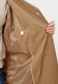Brun kappa inuti med en slät textur, med en ficka som har skötselanvisningar och vit kantning längs kanten. Knappar synliga.