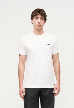 Jeune homme aux cheveux bouclés portant un t-shirt blanc uni à manches courtes avec un petit logo noir sur la poitrine et un pantalon cargo noir.