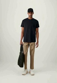 Man wearing black cap, black t-shirt, beige pants, white sneakers, holding black quilted tote bag, standing against plain light background.