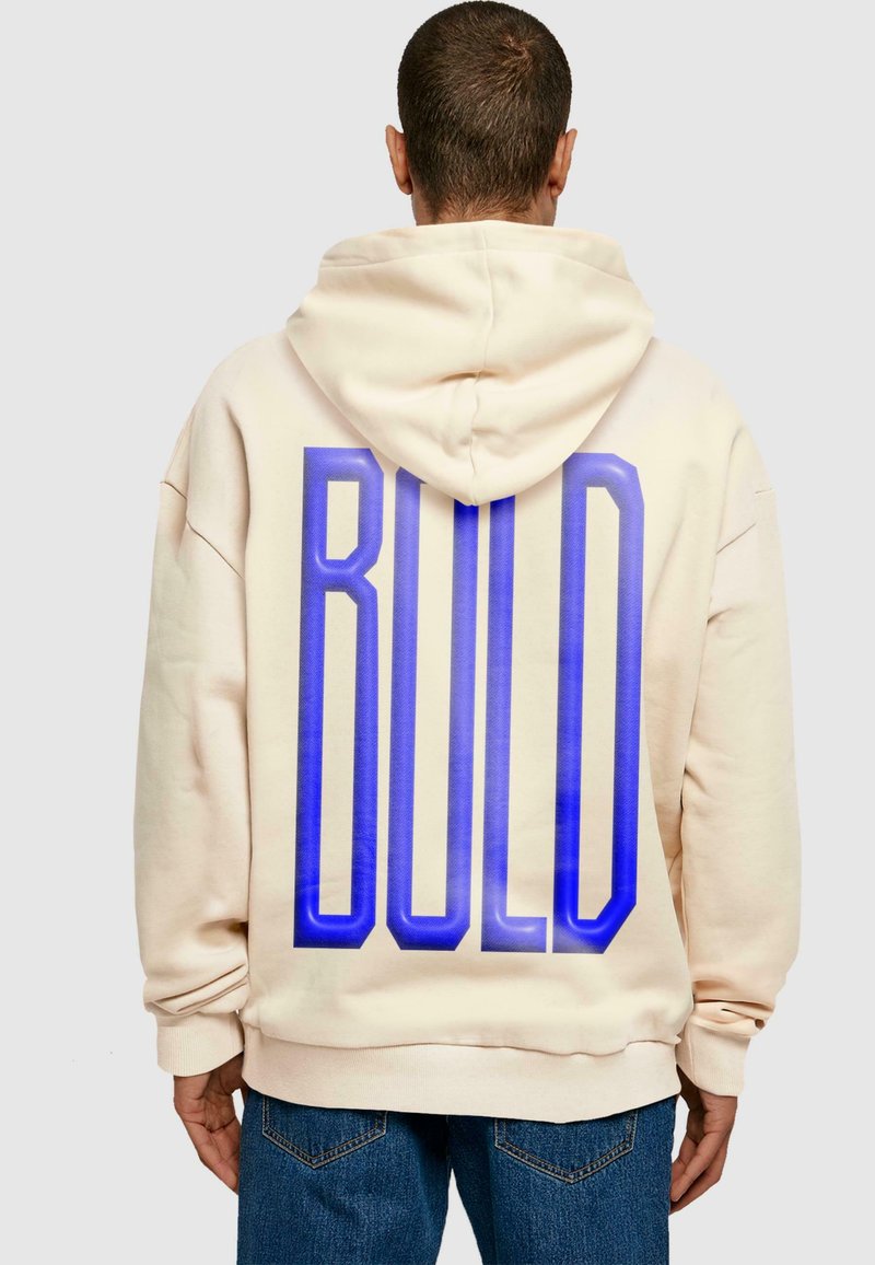 Beige, oversize kapucnis felső, duplán bélelt kapucnival. Nagy, kék "BOLD" felirat található a hátsó részén. Puha, texturált anyagból készült.
