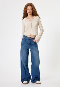 Beige kurz geschnittenes Hemd mit Knöpfen und langen Ärmeln, kombiniert mit weit geschnittenen blauen Jeans und weißen Sneakers. Glatte Textur, lässige Passform.