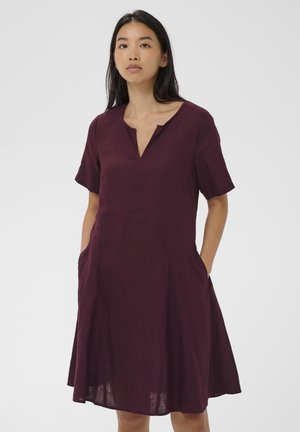 Jauna moteris vilki trumpomis rankovėmis, kelius siekiančią slyvų spalvos suknelę su išpjauta iškirpte ir rankomis kišenėse, stovinti priešais paprastą foną.