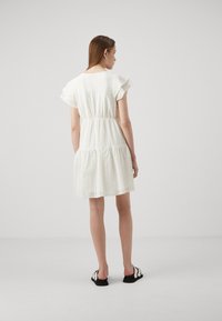 White lace dress with short ruffled sleeves, tiered design, and an elastic waist. Model wearing flat black shoes with white accents.