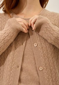 Beige gestrickte Strickjacke mit Kabelmuster, Rundhalsausschnitt, langen Ärmeln und zwei hellen Knöpfen vorne.