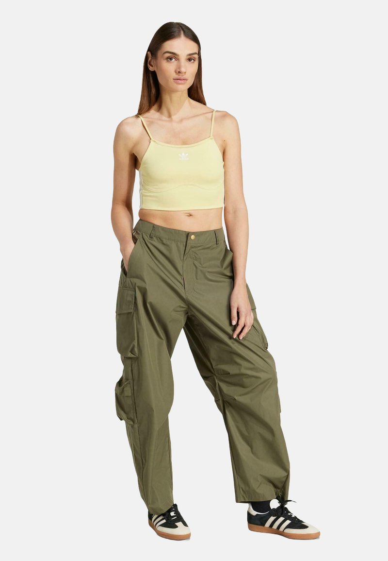 Femme portant un débardeur cropped jaune clair, un pantalon cargo vert olive et des baskets Adidas noir et blanc, se tenant avec les mains dans les poches.