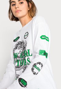 T-shirt de manga longa branca com um design gráfico de dragão em preto, com texto verde e remendos nas mangas, feito de um tecido de algodão macio.
