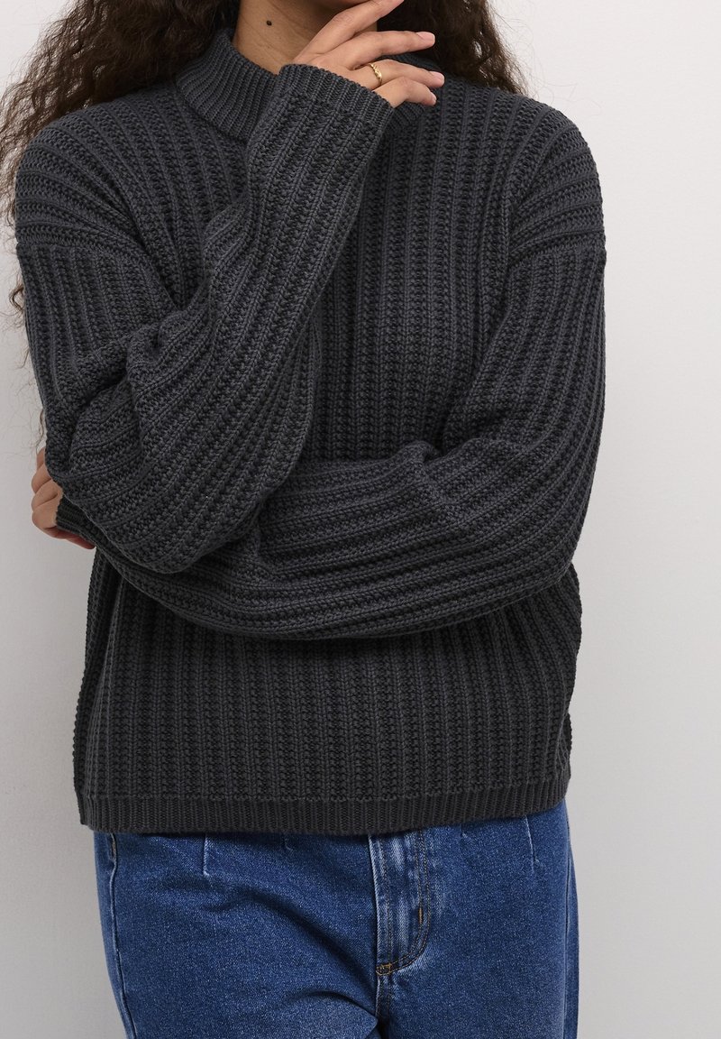 Personne portant un pull en maille côtelée gris foncé et un jean bleu, avec une main touchant le menton et l'autre bras croisé.