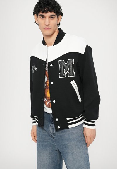 Čierno-biela univerzitná bunda s veľkým emblémom "M", pruhovanými manžetami a bočnými vreckami. Nosená cez tričko s potlačou a modré džínsy.
