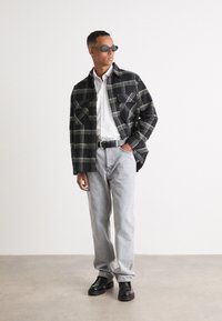 Svart-hvitt rutete jakke med to brystlommer, hvit skjorte, lys grå jeans og svarte platåsko. Modell som bruker solbriller.