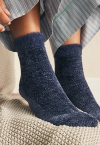 Navyblå, bløde sokker med en blød struktur og en fyldig pelskant, placeret på et tekstureret beige tæppe. Synligt hudområde over sokkerne.