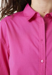 Camisa rosa de botones con cuello clásico, hecha de una tela suave. Presenta un cierre frontal con botones y un ligero brillo.