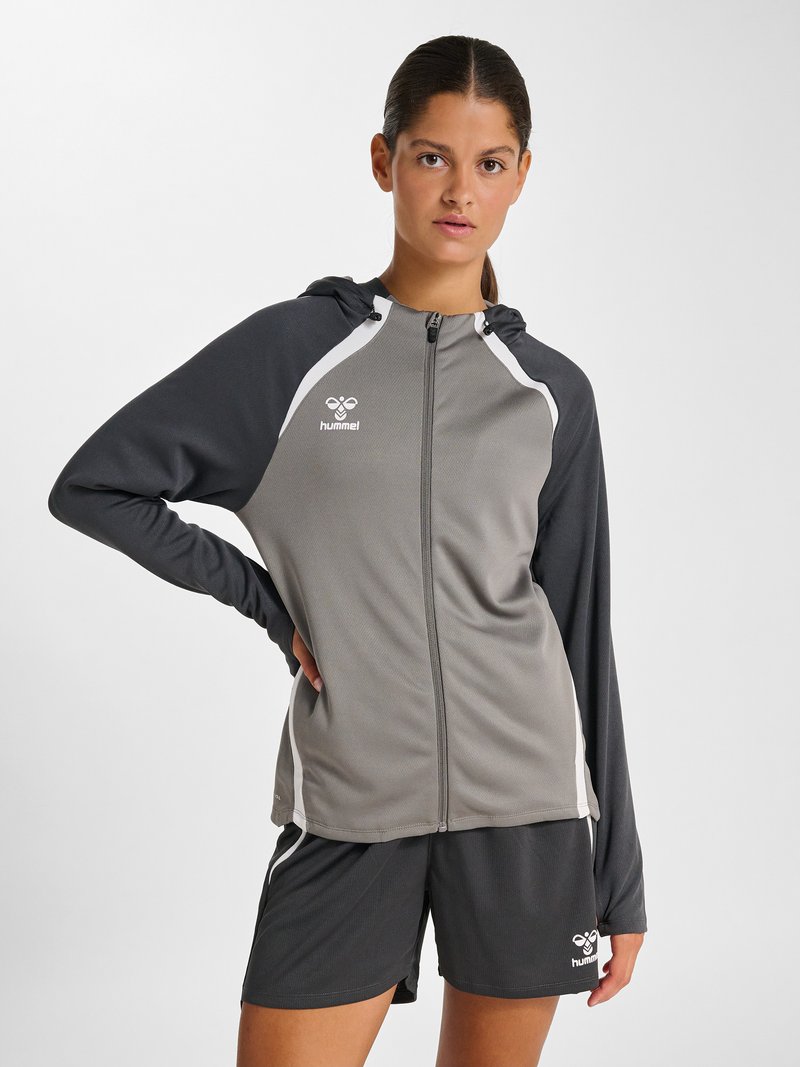 Mujer con sudadera deportiva gris con cremallera y pantalones cortos negros, de pie con una mano en la cadera, expresión neutral, fondo claro y liso.