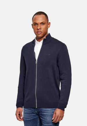 Hombre con un suéter azul marino con cremallera encima de una camisa blanca y vaqueros azules, posando frente a un fondo claro y liso.