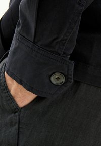 Camicia blu scuro con polsini abbottonati, caratterizzata da una finitura testurizzata. La camicia è abbinata a pantaloni grigio scuro visibili nell'immagine.