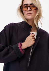 Donkerpaars stoffen jasje met een hoge kraag, voorzien van een getextureerde manchetten in zwart en roze patroon. Draagt oversized schildpadbril.