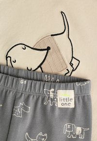 Baumwolloutfit für Kleinkinder mit einem cremefarbenen Oberteil, auf dem ein gestickter Hund abgebildet ist, und einer grauen Shorts mit weißen Hundemustern. Das Etikett trägt die Aufschrift "Hallo kleiner Schatz".