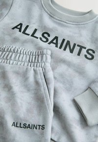 Šviesiai pilka sportinė palaidinė ir kelnės su juodu užrašu "ALLSAINTS". Palaidinė turi briaunotą apykaklę, o kelnės – elastingą juosmenį.