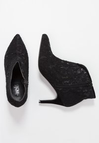 Bottines noires en dentelle avec une pointe aiguë, un design à découpe basse, une fermeture éclair sur le côté et un talon stiletto fin. Douce texture textile et dentelle.
