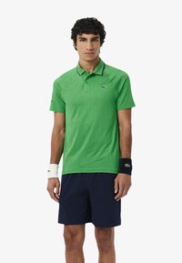 Polo verde con un patrón texturizado y detalles en azul marino, combinado con pantalones cortos azul marino; el modelo lleva pulseras blancas y azul marino.