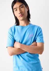 Svetle modré tričko s krátkým rukávem vyrobené z bavlny, s růžovým textem "SCOTCH & SODA" s designem kapek na levé hrudi.
