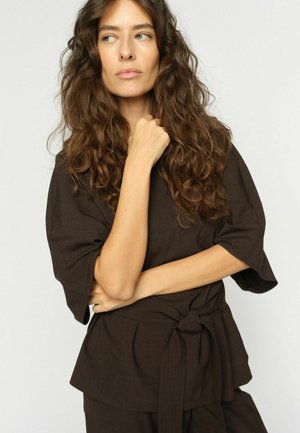 Žena s dlhými kučeravými hnedými vlasmi, oblečená v tmavohnedom krátkym rukávom tričku s opaskom na zaviazanie, sa pozerá doľava s jednou rukou pri brade.