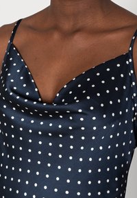 Robe satin bleu marine avec des pois blancs, dotée de bretelles fines et d'un décolleté drapé sur une personne à la peau foncée.