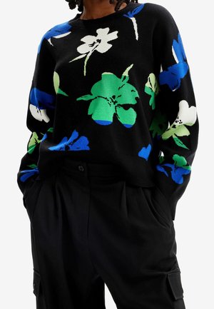 Suéter negro con grandes patrones florales en azul, verde y blanco, combinado con pantalones negros, manos en los bolsillos.