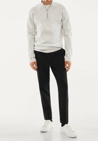 Homme portant un pull gris clair à fermeture éclair quart, pantalon noir ajusté et baskets blanches, debout devant un fond uni.