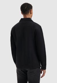 Zwarte wollen shirtjacket met kraag, knoopmanchetten en een gestructureerde stof, voorzien van een rechte zoom en minimale naden aan de achterkant.