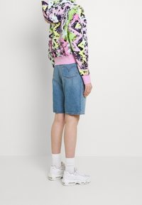 Felpa con cappuccio dal motivo astratto colorato in rosa, verde e nero; abbinata a pantaloni shorts in denim azzurro chiaro e sneakers bianche.