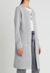 Šedý pletený kardigan s žebrovanou strukturou, otevřeným předním dílem, dlouhými rukávy a dvěma předními kapsami, kombinovaný s bílým tričkem a džínami.