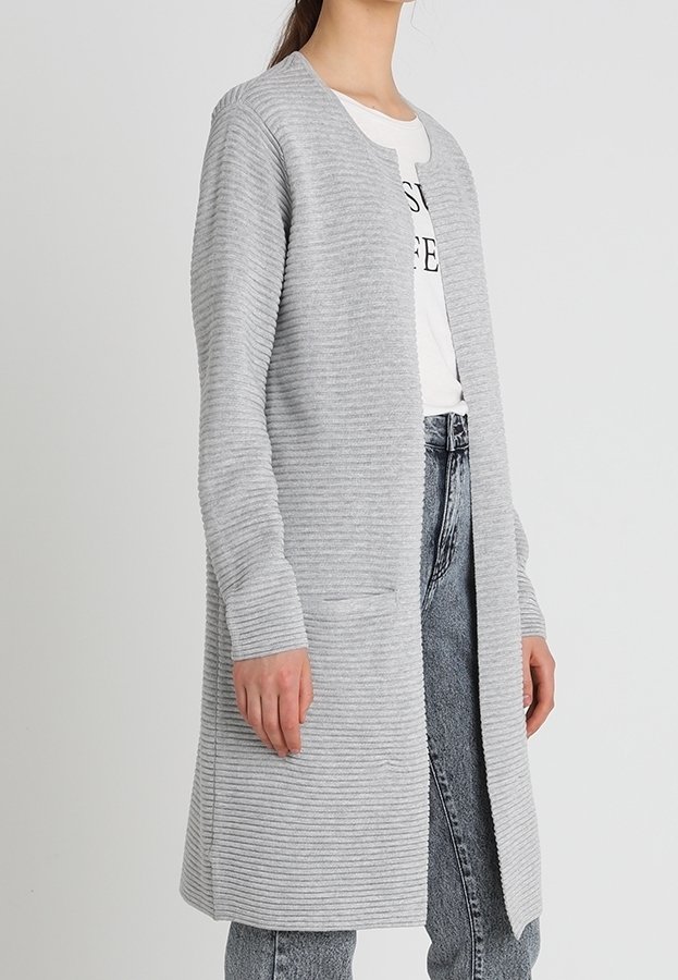Šedý pletený kardigan s žebrovanou strukturou, otevřeným předním dílem, dlouhými rukávy a dvěma předními kapsami, kombinovaný s bílým tričkem a džínami.