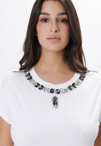 Camiseta blanca con un cuello decorativo de piedras de rhinestone negras y transparentes, con un acento de gema central debajo del escote. Textura de tela suave.