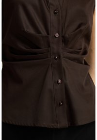 Braune blusige Bluse mit gefalteten Details, verziert mit glänzenden Knöpfen und einem glatten, leichten Stoff. Das Design umfasst vertikale Nähte und eine taillierte Passform.