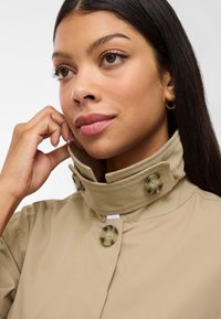 Cappotto beige con un colletto alto, due grandi bottoni decorativi e una texture liscia. La modella tocca il colletto, evidenziando i dettagli.