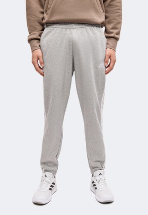Άτομο ντυμένο με ανοιχτό γκρι φόρμα Adidas, μπεζ φούτερ και λευκά αθλητικά παπούτσια Adidas, στέκεται μπροστά σε λευκό φόντο.