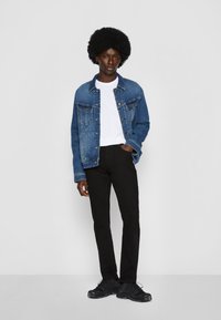 Persona de pie con una mano en el bolsillo, vistiendo una chaqueta de mezclilla azul, camiseta blanca, pantalones negros y zapatillas negras, frente a un fondo liso.