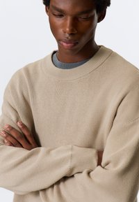 Μπεζ πλεκτό πουλόβερ με άνετη γραμμή, ριμπ λαιμόκοψη και απαλή υφή. Τα μανίκια είναι ελαφρώς φαρδιά, προσδίδοντας άνεση και ευκολία.