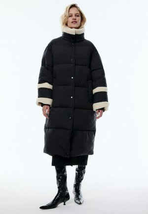 Schwarze Daunenjacke mit hohem Kragen und cremefarbenem Kunstpelzbesatz. Verfügt über große gepolsterte Abschnitte und Knopfverschlüsse. Wird über schwarzer Kleidung und glänzenden Stiefeln getragen.