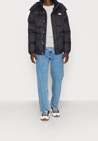 Giubbotto puffer nero con cappuccio, tasche frontali e bottoni a pressione, indossato sopra un maglione grigio e jeans azzurri chiari con sneakers bianche.