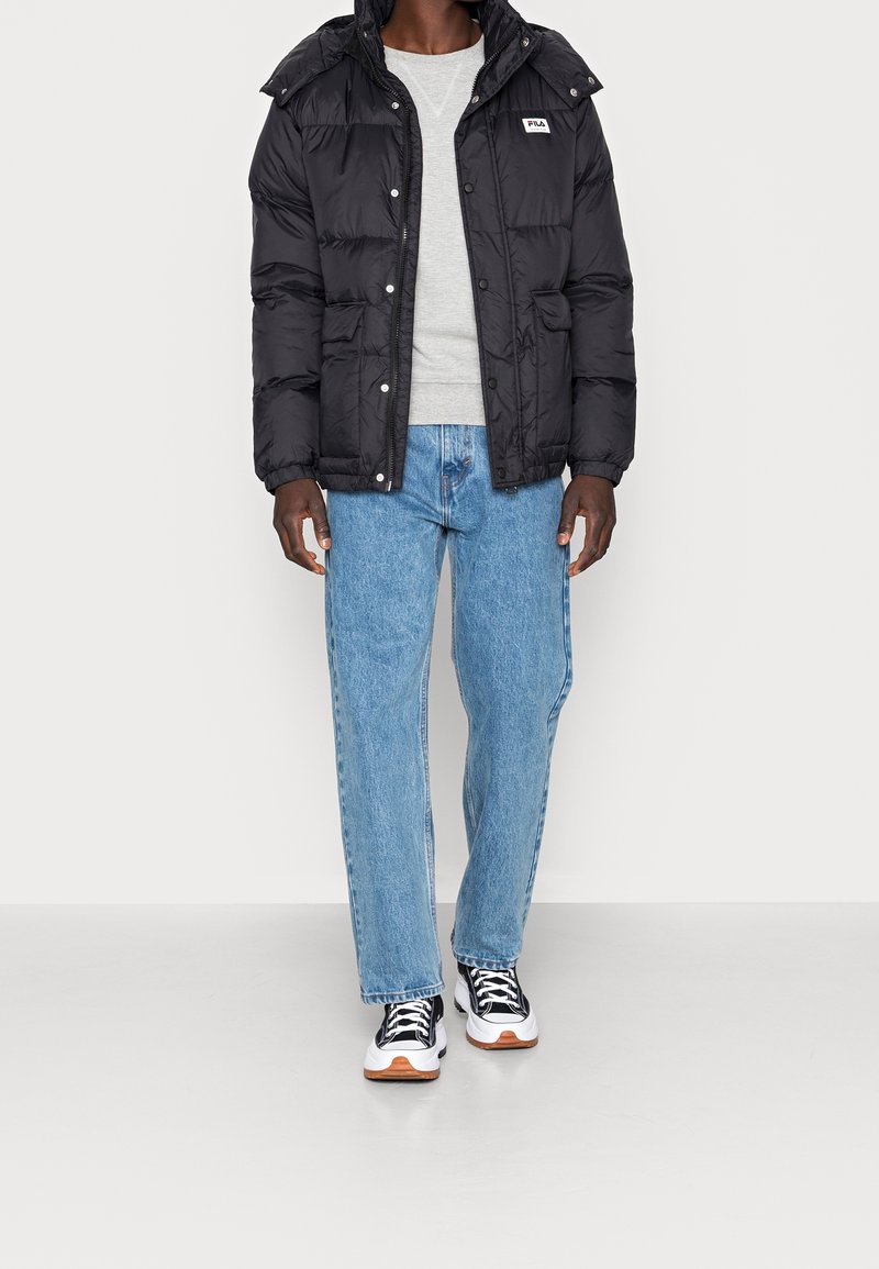 Giubbotto puffer nero con cappuccio, tasche frontali e bottoni a pressione, indossato sopra un maglione grigio e jeans azzurri chiari con sneakers bianche.