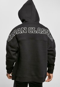Čierna mikina s kapucňou s veľkým potlačou "URBAN CLASSICS" na chrbte. Má kapucňu a rebrované manžety, vyrobená z mäkkej, štruktúrovanej tkaniny.