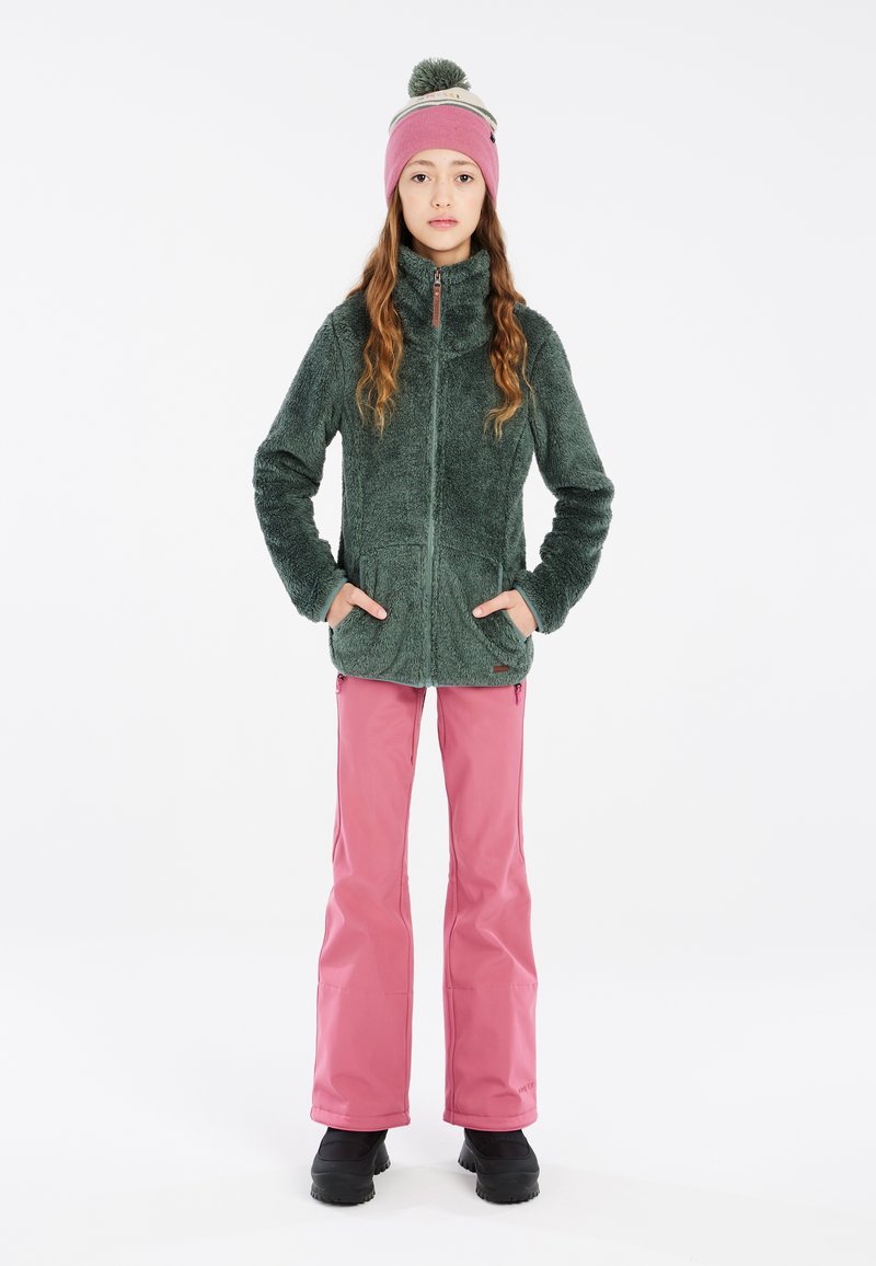 Giacca in pile verde con colletto alto, chiusura con zip e tasche laterali. Abbinata a pantaloni impermeabili rosa e stivali neri.