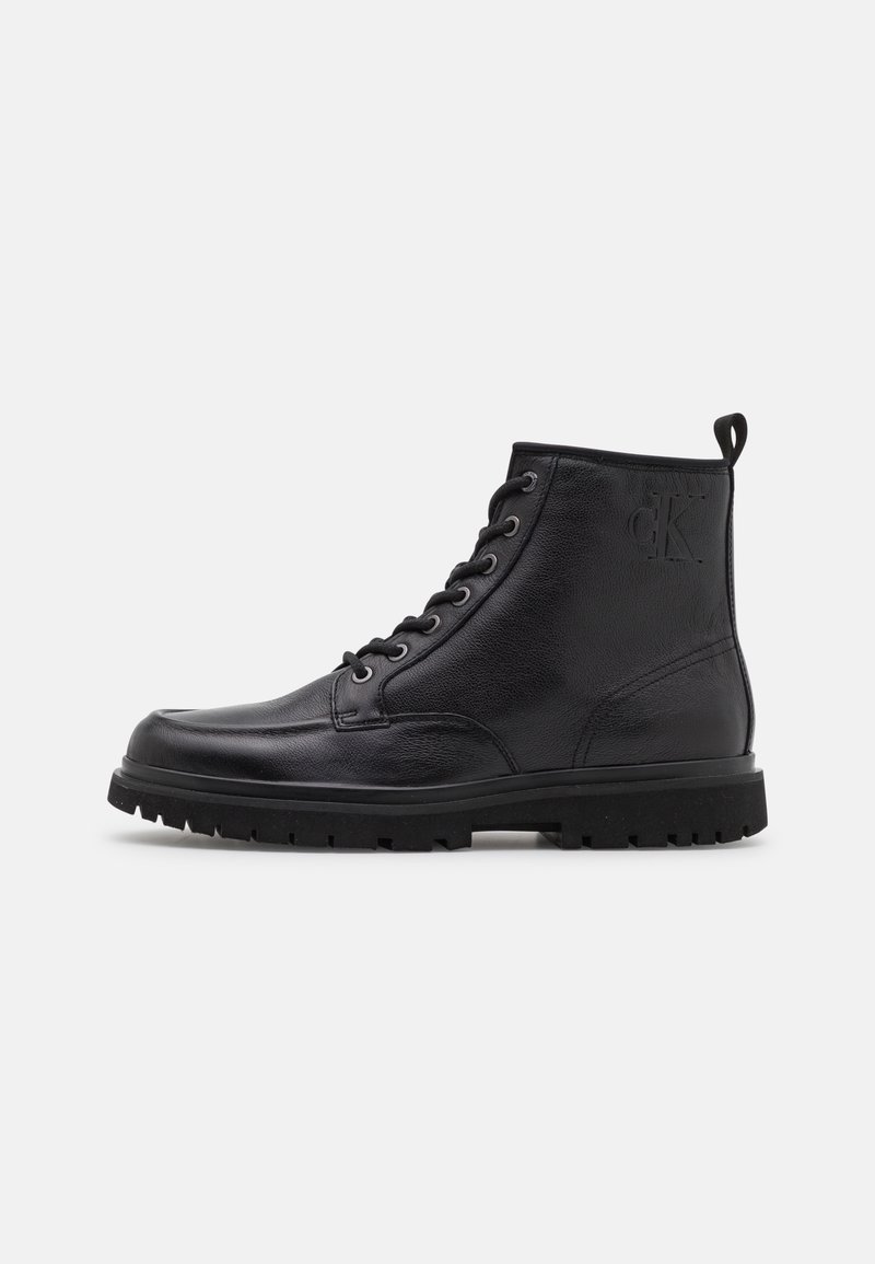 Bottine en cuir noir avec design à lacets, surface texturée, bout rond, et une semelle en caoutchouc avec un motif de bande de roulement. Logo embossé sur le côté.