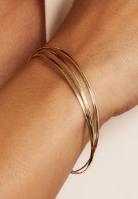 Bracelets en métal doré, trois fins et lisses, reposant sur une surface de peau texturée. Design simple et élégant, finition réfléchissante.